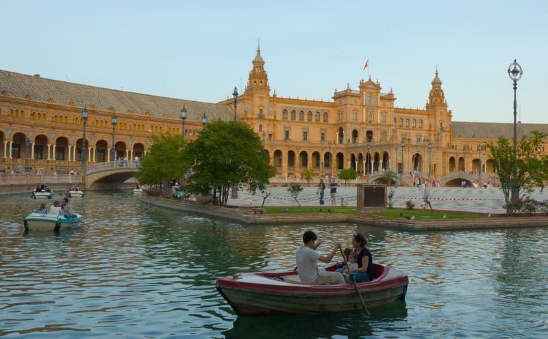 Casal remando num barco no canal da Plaza de España, em Sevilha, com o edifício de tijolo e as torres ao fundo